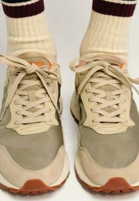 Zapatillas beige con paneles en tono marrón y oliva, con puntera redonda, cordones y suela texturizada. Llevadas con calcetines crema con rayas color burdeos.