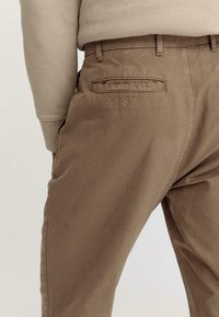 Pantaloni in cotone marroni con una texture liscia, dotati di tasca posteriore e passanti per la cintura. La vita è aderente e sono visibili tasche laterali.
