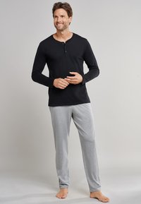 Schwarz, langärmliges Henley-Top mit Knopfleiste, kombiniert mit grauen Pyjamahosen aus einem weichen, leichten Stoff. Barfußhaltung.
