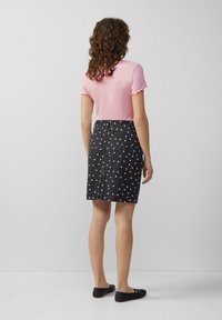 Eine Frau mit lockigem Haar steht vor einer grauen Wand. Sie trägt ein rosa Kurzarmoberteil, einen schwarzen Rock mit weißen Punkten und schwarze Ballerinas.