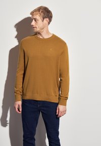 Senfgelbes Strickpullover mit Rundhalsausschnitt, langen Ärmeln, gerippten Bündchen und Saum, mit einem kleinen Logo auf der Brust. Kombiniert mit dunklen Jeans.