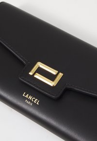 Svart lær lommebok med en gullfarget lås med geometrisk design og preget "LANCEL PARIS"-logo i et nedre hjørne.