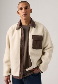 Chaqueta de forro polar color crema con un cuello marrón y un bolsillo de parche; presenta un cierre de cremallera en la parte delantera y tejido texturizado. Usada sobre una camiseta gris.