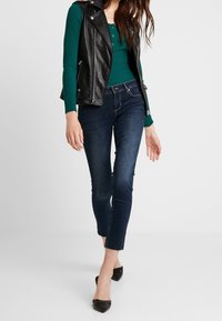 Gilet en cuir noir sur un haut à manches longues verde côtelé, associé à un jean slim bleu foncé et des escarpins noirs à bout pointu.