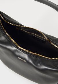 Geantă de piele neagră cu fermoar aurit, căptușeala interioară vizibilă, branduită cu "Calvin Klein." Textură netedă, formă rotundă, un singur mâner.