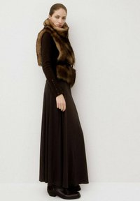 Estola de piel marrón llevada sobre un suéter negro ajustado y un vestido maxi negro fluido. Presenta mangas largas y detalles de botones. Botas negras hasta el tobillo.