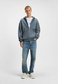 Grijze zip-up hoodie, katoenmix stof, voorzakken, gedragen met witte T-shirt, versleten lichtblauwe jeans en beige sneakers.