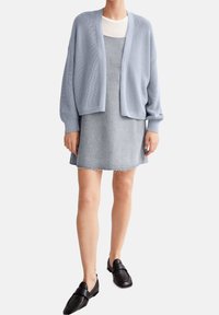 Vaaleansininen ribbineulottu neuletakki harmaan, tekstuurisen mekon päällä, jossa on rispaantunut helma. Mustat loafersit täydentävät asun.