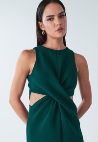 Zelena zvita obleka z teksturirano površino, z izrezanimi stranicami in visokim ovratnikom. Enostavna zasnova brez dodatnih vzorcev ali poudarkov.
