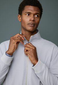 Man with short curly hair zips up a light gray jacket, looking to the side against a plain gray background.