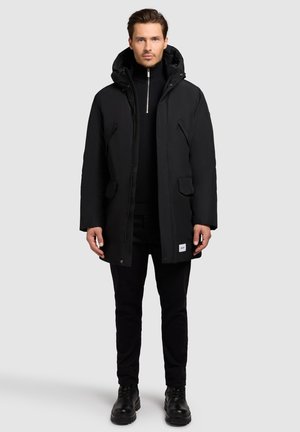 Sort isoleret parka-jakke med hætte, lodret frontlynlås, sidelommer og et logo-patch i kanten. Båret over en sort turtleneck.