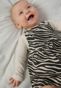 En baby har på sig en beige långärmad tröja under en svart- och beige randig overall-klänning med knappar och en ficka.