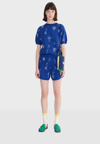 Femme portant un ensemble assorti bleu avec des imprimés botaniques blancs, des chaussettes blanches à rayures jaunes, des chaussures vertes, et un petit sac à épaule bleu.