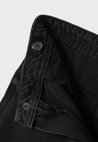 Črna denim tkanina z vidnim zapiranjem na gumb in pasom. Ima teksturirano površino in rahlo franže na robovih.