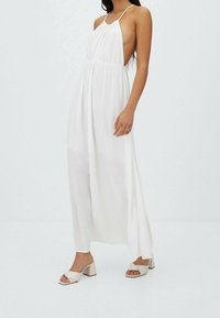 Weißes Maxikleid mit Neckholder-Ausschnitt, seitlichen Cutouts und fließendem Stoff. Kombiniert mit hellbeigen Sandalen mit Blockabsatz und Schleifendetail.