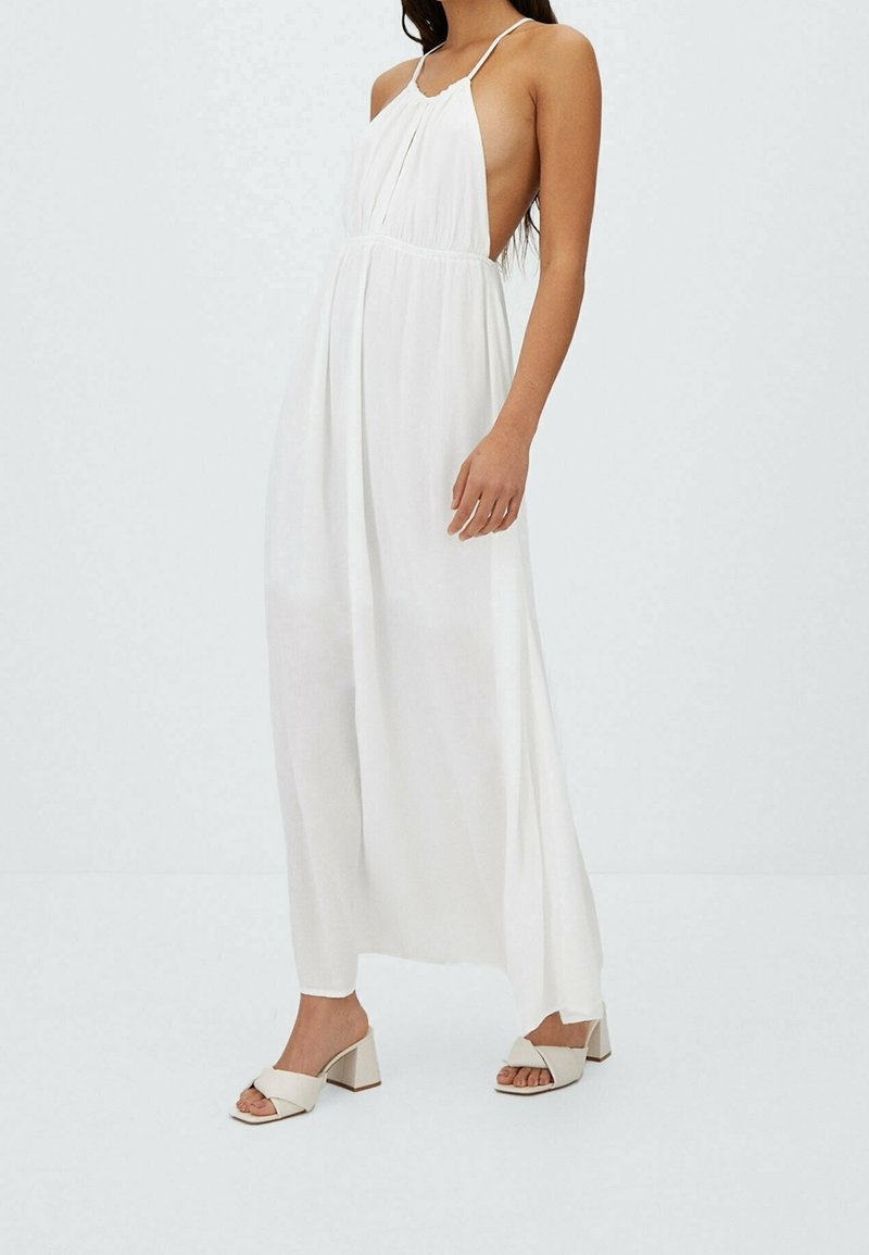 Weißes Maxikleid mit Neckholder-Ausschnitt, seitlichen Cutouts und fließendem Stoff. Kombiniert mit hellbeigen Sandalen mit Blockabsatz und Schleifendetail.