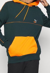 Zielona bluza z kapturem z pomarańczowymi akcentami i sznurkami. Posiada kieszeń typu kangur i małe logo na klatce piersiowej. Materiał wydaje się miękki i strukturalny.