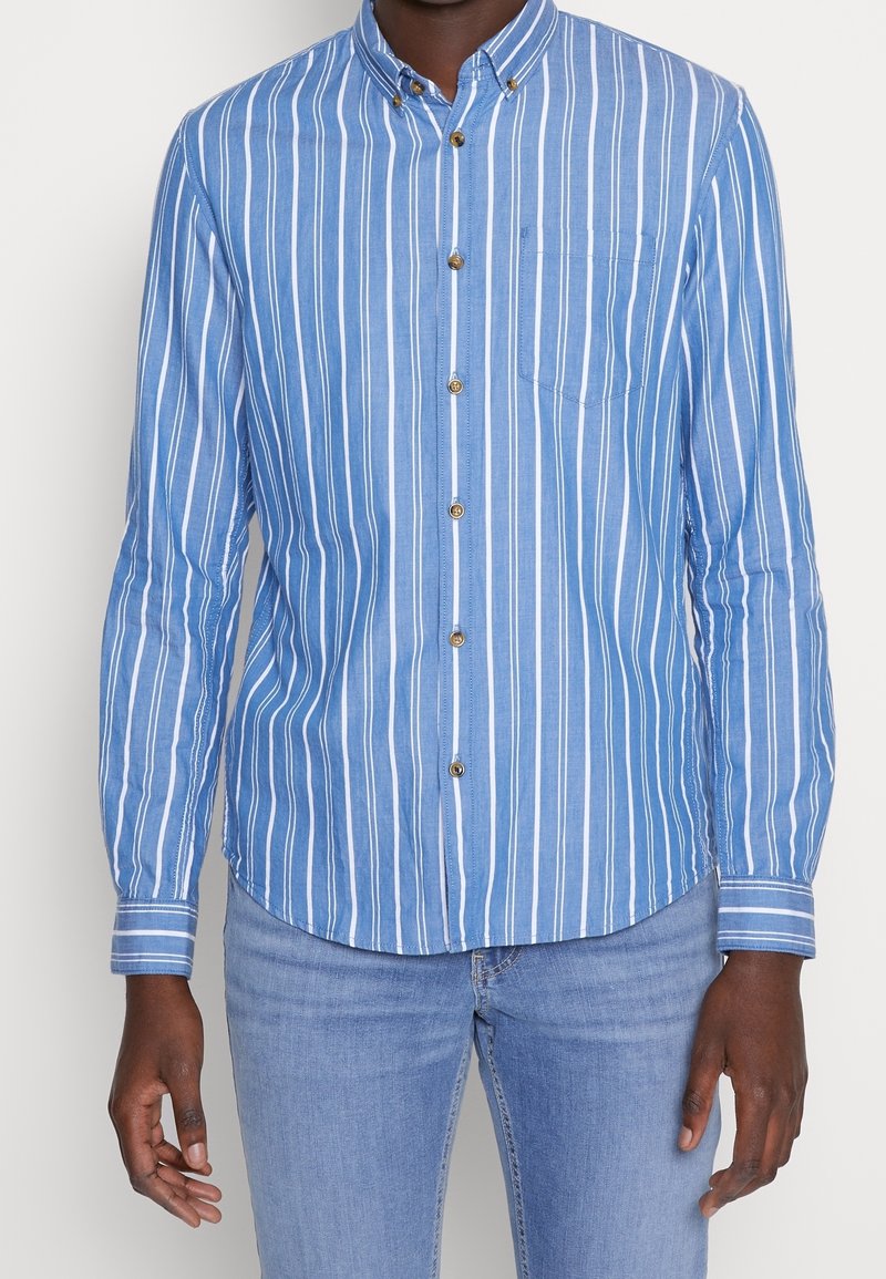 Homme portant une chemise à rayures verticales bleues et blanches avec une poche poitrine et un jean bleu clair, debout devant un fond uni.