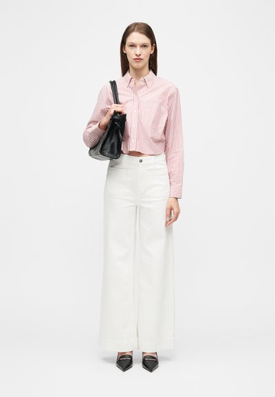 Femme portant un haut court rayé rose et blanc, un pantalon blanc à jambes larges, des chaussures noires pointues, et tenant un sac à bandoulière noir.