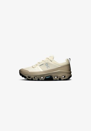 Beiger und hellbrauner Schnür-Sportschuh mit dicker, robuster schwarzer Sohle, entworfen für Outdoor-Trailrunning oder Wandern.