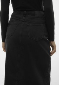 Černá džínová sukně s vysokým pasem, zadními kapsami a rovnou siluetou. Textura vypadá robustně, s minimálními detaily a čistým zakončením.