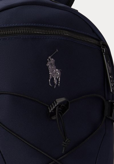 Κοντινή λήψη μπροστινής θήκης σάκιδιου πλάτης σε ναυτικό μπλε με μαύρο φερμουάρ, ελαστικό κορδόνι και κεντημένο λογότυπο παίκτη πόλο πάνω σε άλογο.