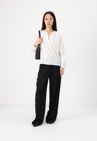Blusa blanca de manga larga con escote en V y un delicado patrón bordado; combinada con pantalones negros holgados; persona sosteniendo un bolso negro.
