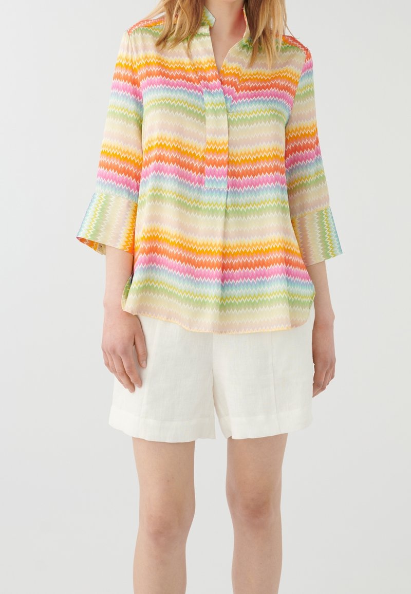 Femme portant un chemisier à manches longues à motif zigzag multicolore et un short blanc longueur genoux, debout devant un fond uni.