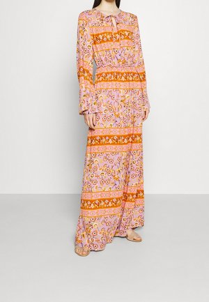 Femme portant une longue robe rose à motifs floraux avec des rayures horizontales orange et des manches longues, debout devant un fond blanc uni.