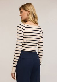Donna con capelli biondi che indossa una maglietta a maniche lunghe a strisce crema e blu navy e pantaloni blu navy a vita alta, rivolta di lato contro uno sfondo semplice.