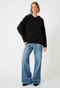 Schwarzer Kapuzenpullover mit Perlenverzierungen, Kängurutasche und gerippten Bündchen, kombiniert mit hellblauen weit geschnittenen Jeans.