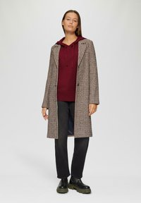 Gekruist lange jas met een getailleerde pasvorm, grijze en bruine tinten, over een bordeauxrode hoodie. Gecombineerd met zwarte jeans en stevige zwarte laarzen.