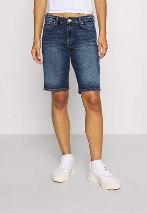 Person iført mørkeblå denimshorts til knæet, hvide sneakers og en kort hvid top, stående på en ensfarvet lysegrå gulv.