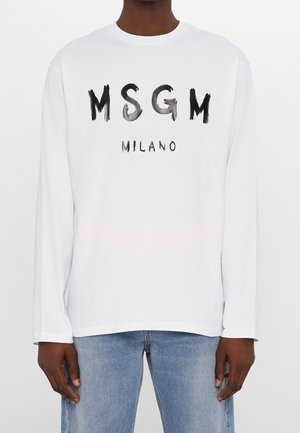 Biała koszula z długim rękawem z czarnym, malowanym stylem napisu "MSGM MILANO", noszona przez osobę w średnio niebieskich dżinsach, stojącą na gładkim białym tle.