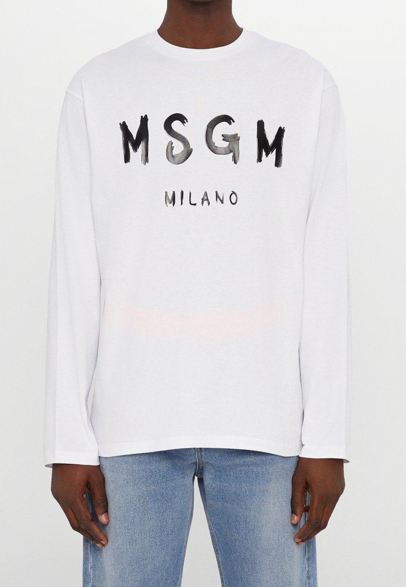 Chemise blanche à manches longues avec texte noir style peinture "MSGM MILANO", portée par une personne portant un jean bleu moyen, debout devant un fond blanc uni.