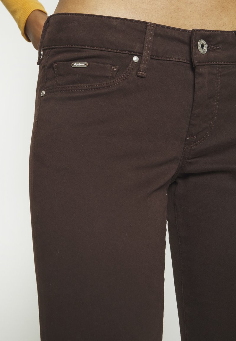 Braune Baumwolljeans mit glatter Textur, ausgestattet mit einer kleinen Vorderasche, einem markierten Metall-Tag und Kontrastnähten.