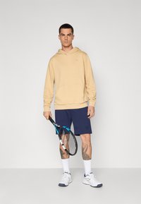 Beige kapucnis pulóver, tengerészkék rövidnadrág, fehér sportcipő, teniszütőt tart a kezében. Kézfejeken tetoválások látszanak, és a rövidnadrágon logó található.