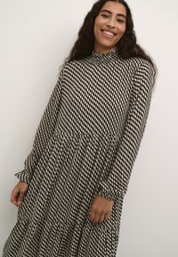 Robe à manches longues noire et crème avec un col montant, des détails smockés et un ourlet à volants en étages. Tissu lisse avec un motif géométrique.