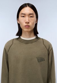 Ungdom med lige, sort hår iført en olivengrøn sweatshirt med rund hals og subtil tekstlogo på brystet, neutral baggrund.