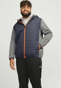 Marineblå puffervest med orange detaljer, grå langærmet skjorte under, sorte bukser. Har hætte og lynlåslommer.