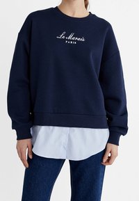 Felpa blu navy con il testo "Le Marais Paris", indossata sopra una camicia button-up a righe blu chiaro e abbinata a jeans blu scuro.