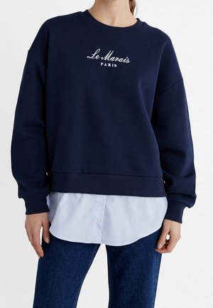 Sudadera azul marino con el texto "Le Marais París", llevada sobre una camisa de botones a rayas azul claro y combinada con unos jeans azul oscuro.