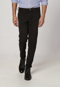 Schwarze Slim-Fit-Hose aus Baumwolle mit einem Knopfverschluss und vorderen Taschen, kombiniert mit schwarzen Schnürstiefeln.