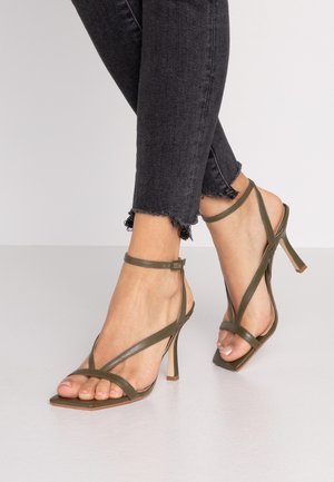 Sandales à talons hauts vert olive avec des brides fines, design à bout carré et boucle au niveau de la cheville. Présentées sur un fond en jean sombre aux ourlets effilochés.