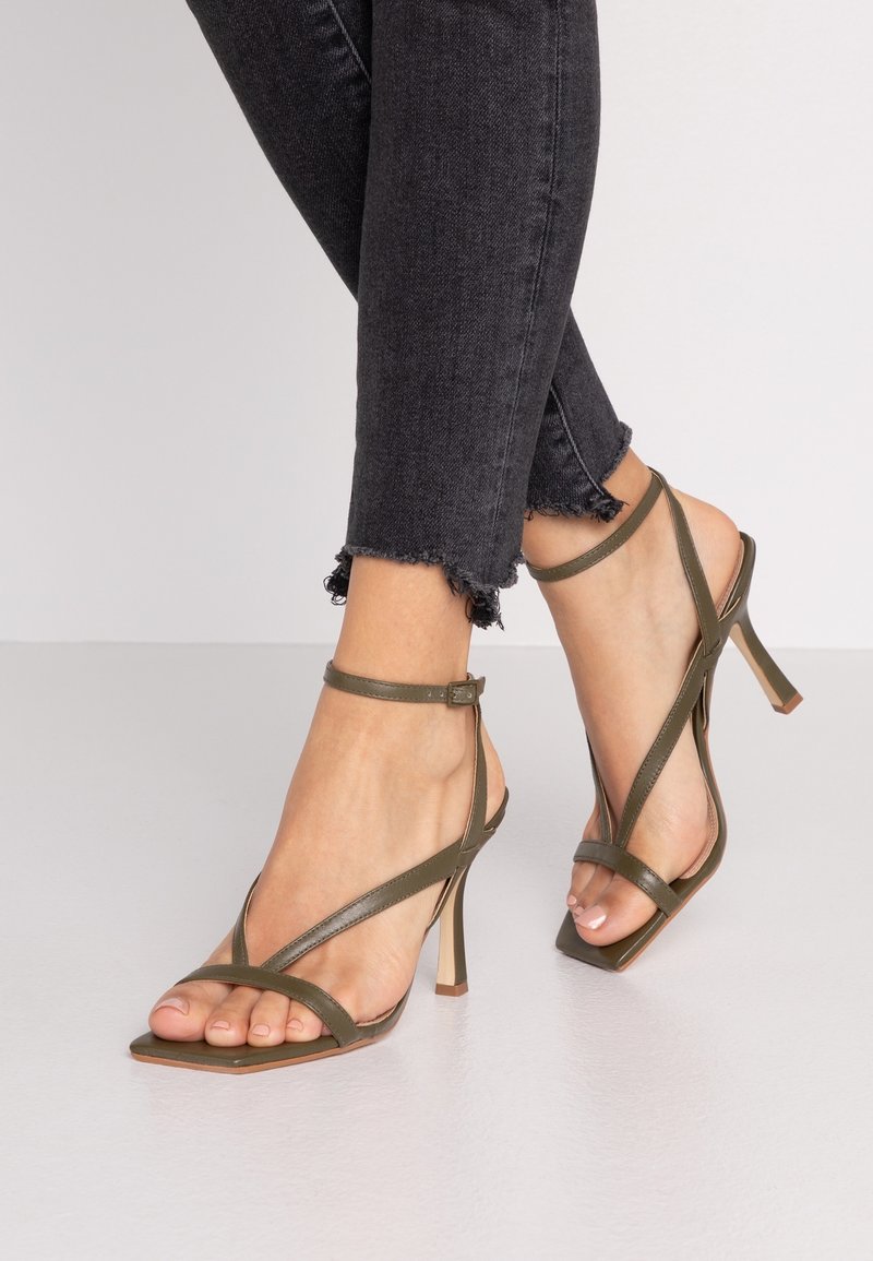 Sandales à talons hauts vert olive avec des brides fines, design à bout carré et boucle au niveau de la cheville. Présentées sur un fond en jean sombre aux ourlets effilochés.