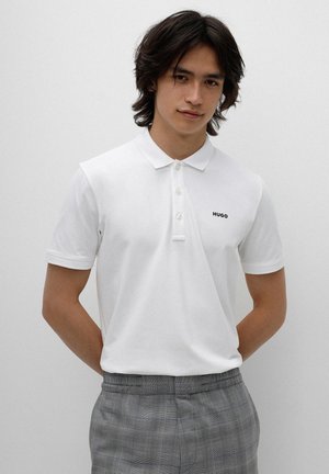 White polo shirt with short sleeves and a three-button placket, featuring a small black "HUGO" logo on the chest. Worn with plaid trousers.