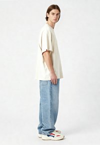 Beige oversize t-paita, siniset väljä istuvuus farkut ja valkoiset tennarit punaisilla ja sinisillä yksityiskohdilla. Kangas näyttää pehmeältä ja teksturoidulta.