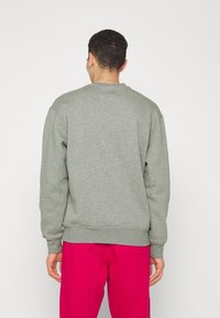 Hombre vestido con un sudadera gris de manga larga y pantalones rojos brillantes, de espaldas a la cámara frente a un fondo blanco liso.