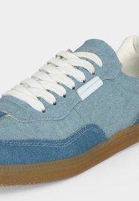 Baskets bleu denim avec lacets blancs, tige en tissu texturé et semelle en caoutchouc naturel, vues en gros plan sous un angle latéral.
