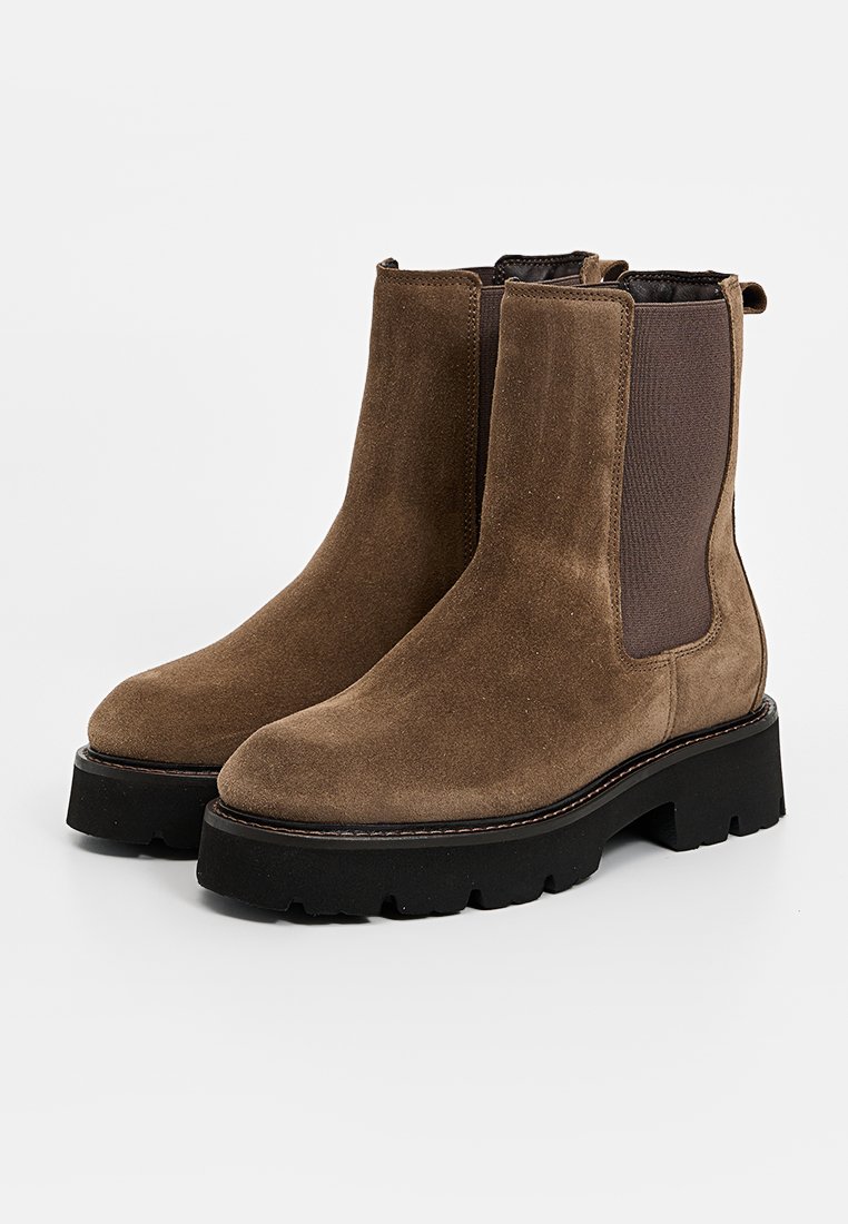 Braune Wildleder-Stiefeletten mit dehnbaren Seiteneinsätzen, einer dicken schwarzen Gummisohle und dekorativen weißen Nähten entlang der Kante.